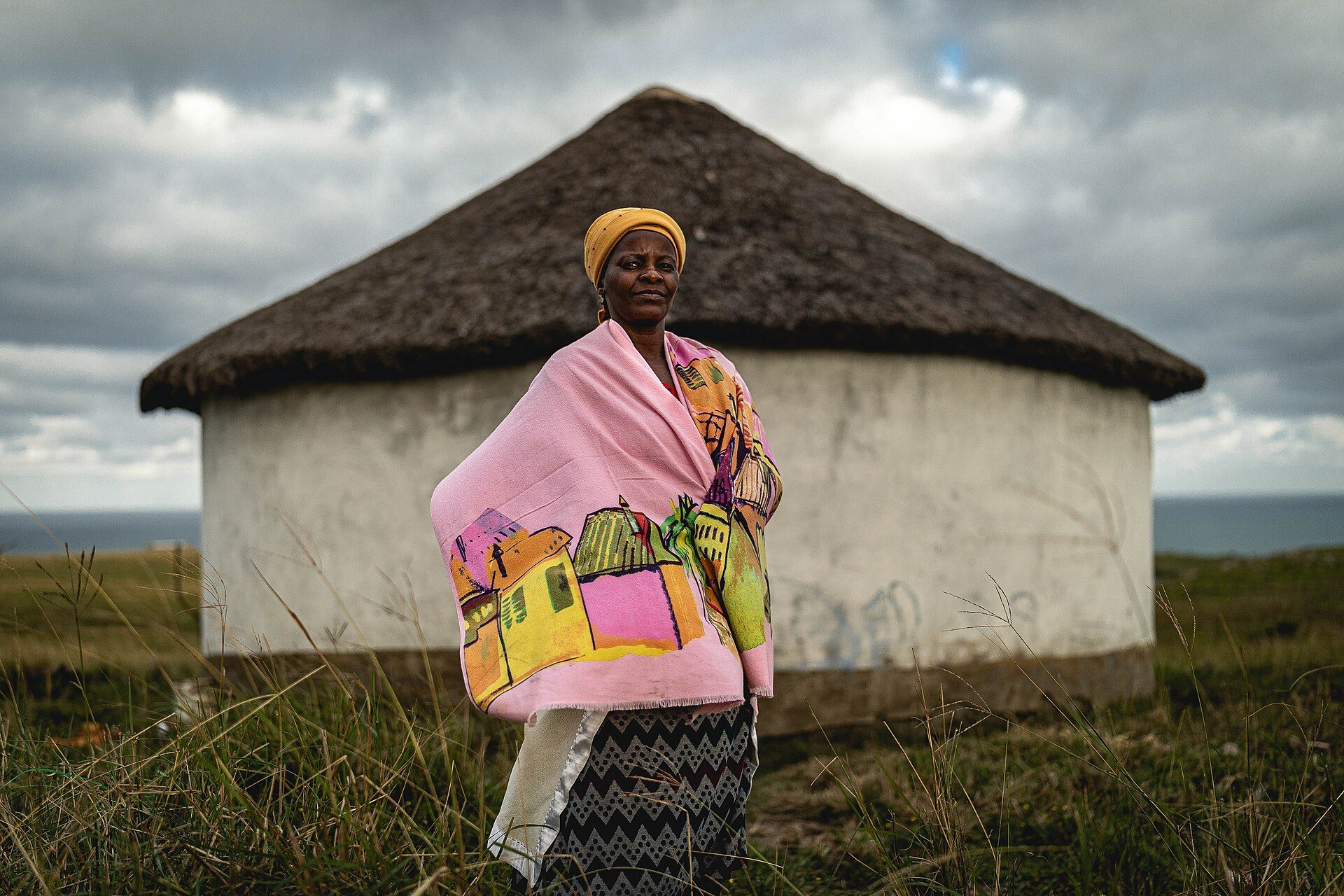 A woman wrapped in a painted blanket stands before a thatched rondavel on the coast, South Africa
