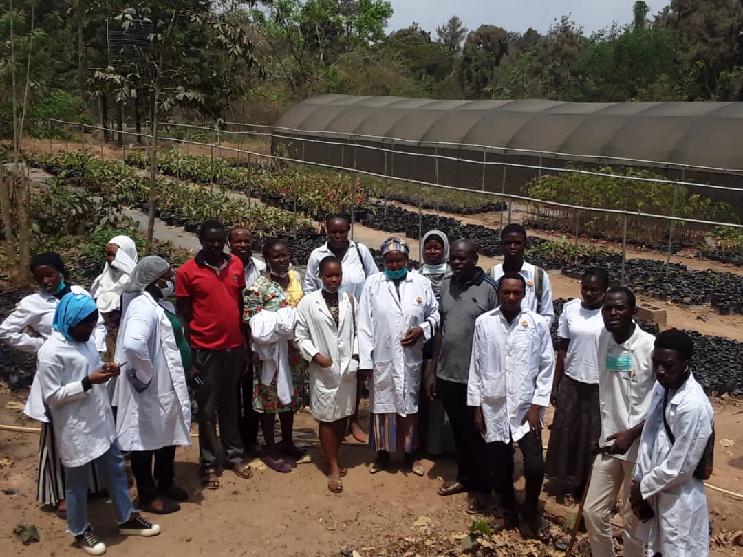 Researchers in white lab coats and community members gathered at a tree improvement nursery beside a greenhouse